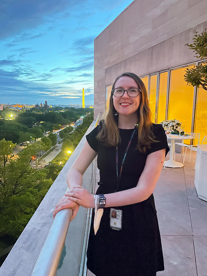 person with long blond hair and glasses in black shirt, standing on a patio with hands on the railing